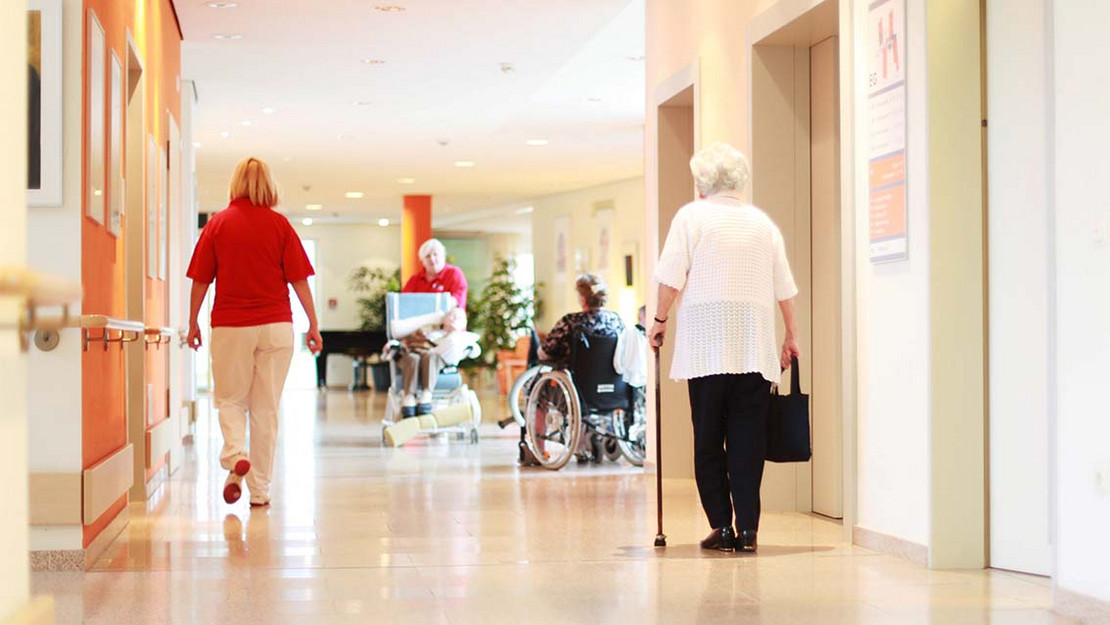 Alte Frau mit einem Gehstock läuft den Flur eines Pflegeheims entlang. Neben ihr eine Pflegekraft.