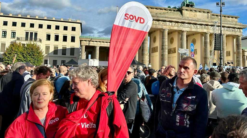 Mann und Frau mit SoVD-Wimpel in einer Menschenmenge vor dem Brandenburger Tor. 