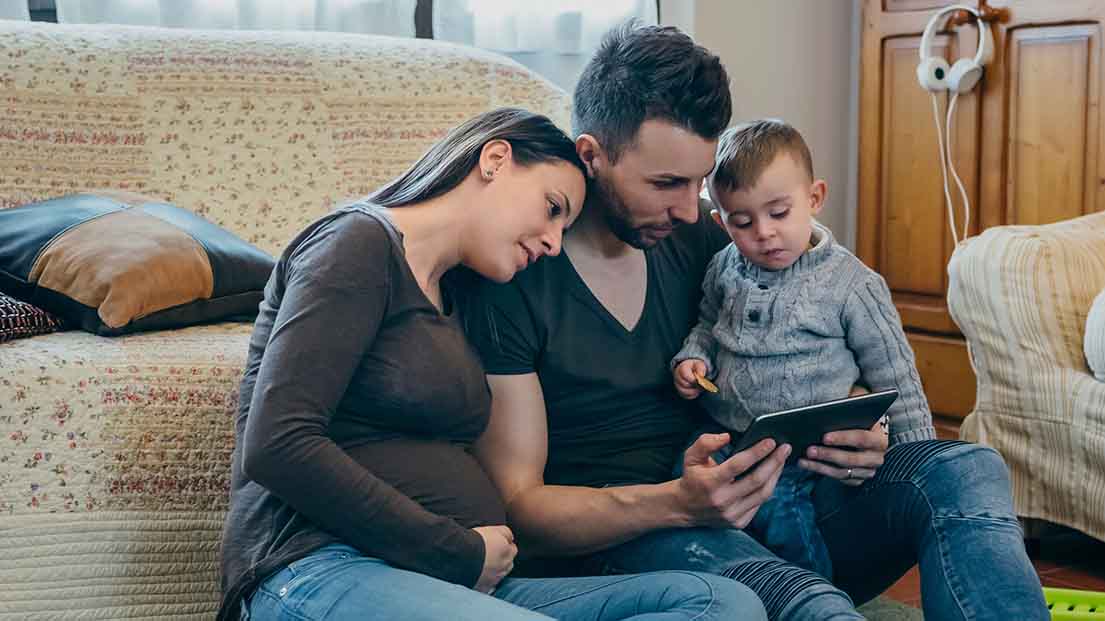 Familie sitzt vor dem Sofa auf dem Boden und schaut in ein Tablet. 