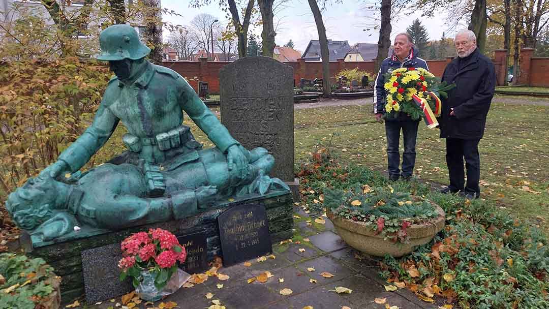 Zwei Männer mit Blumengesteck vor Kriegsgefallenendenkmal
